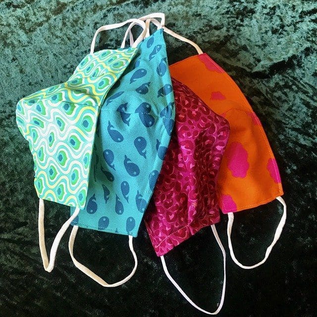 Four colorful fabric face masks with various patterns, including wavy green, blue whales, magenta abstract, and orange with pink spots, displayed on a dark green background.
