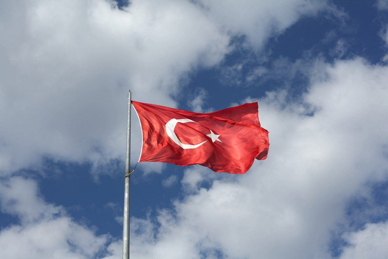The red Turkish flag with a white crescent moon and star waves on a flagpole against a blue sky with white clouds.