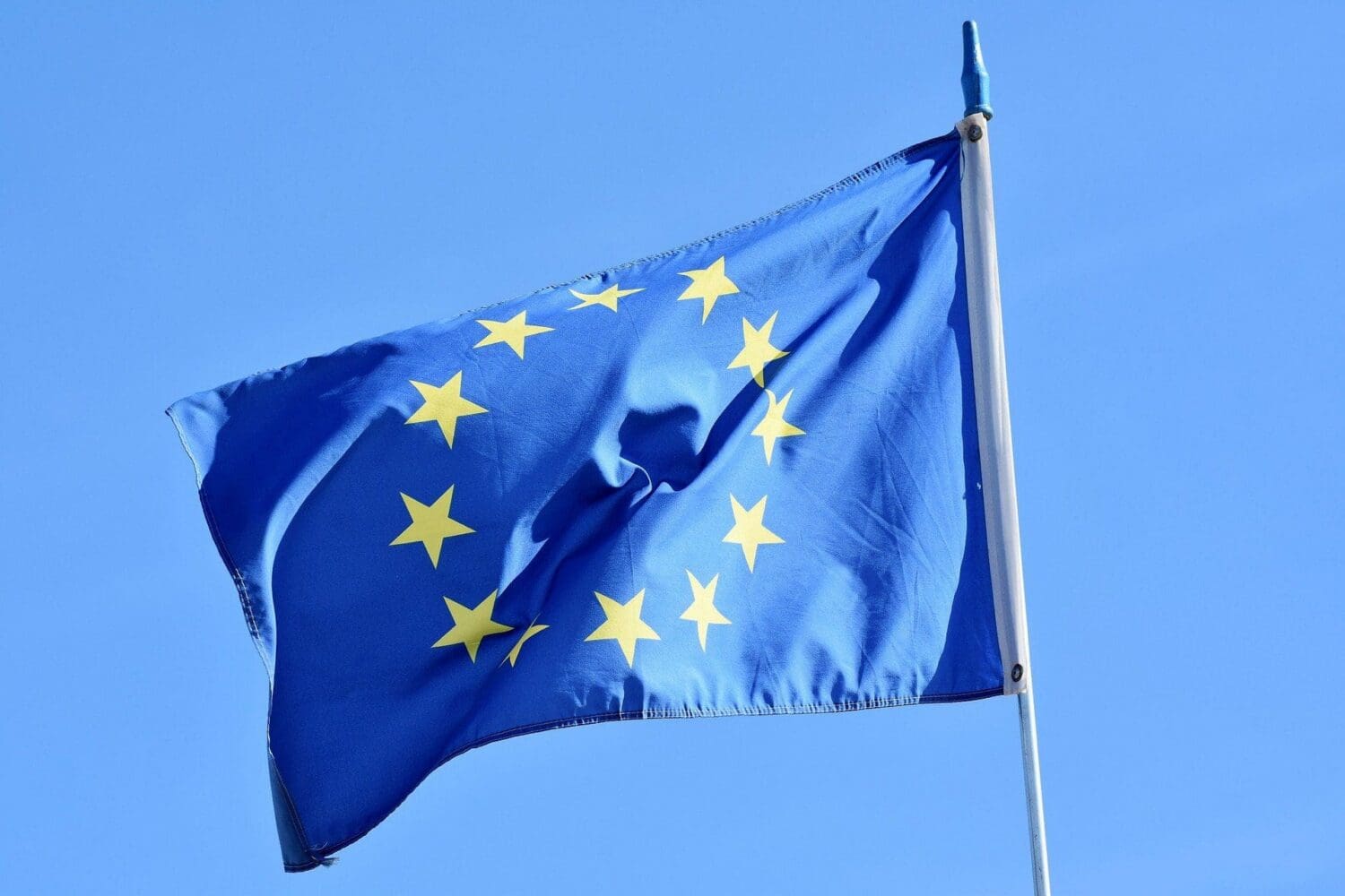 The European Union flag, blue with a circle of twelve golden stars, waving against a clear blue sky.