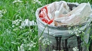 A dark, stained trash can filled with white and red plastic bags, surrounded by green grass and white wildflowers.