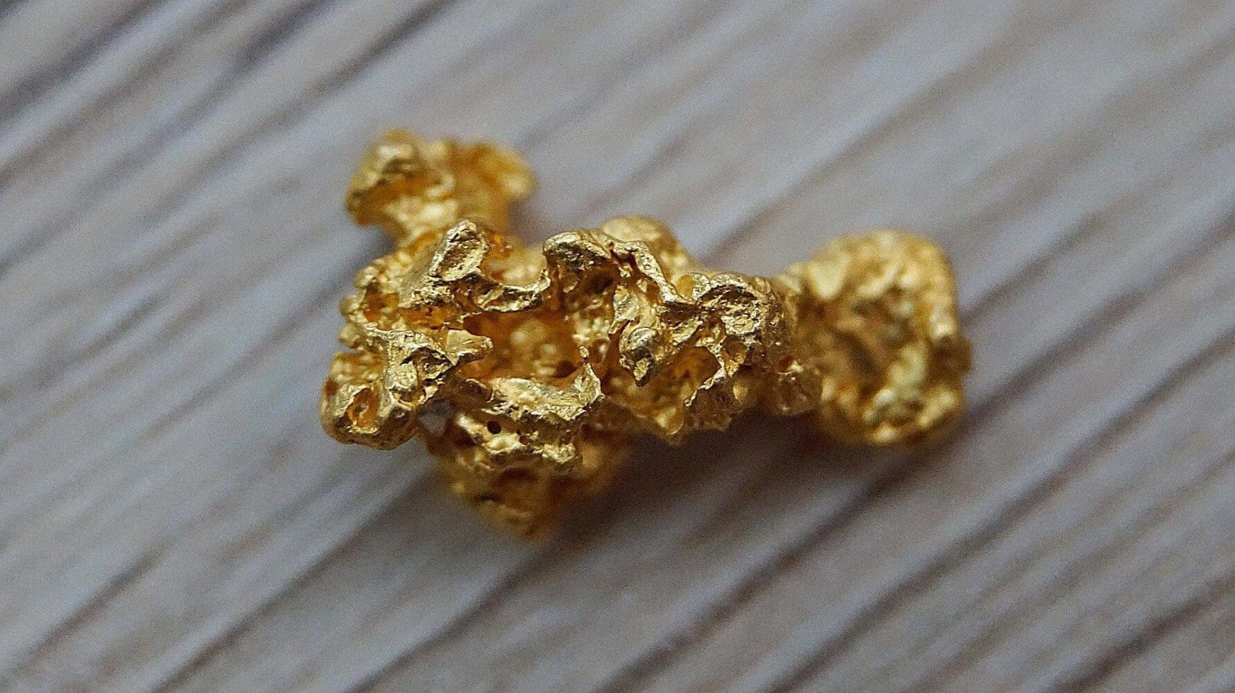 A close-up of a raw, textured gold nugget on a light wooden surface.