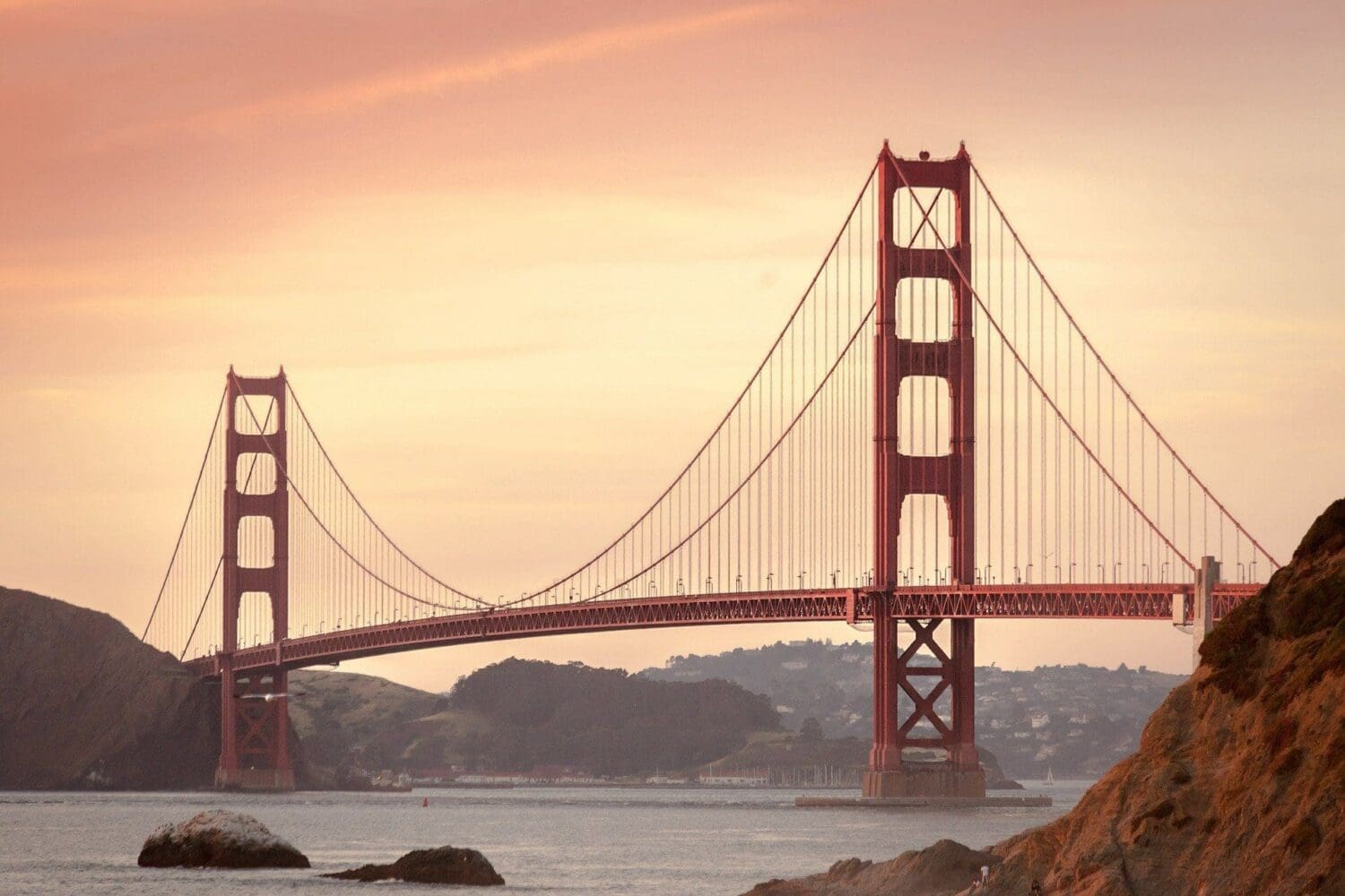 The Golden Gate Bridge, a red-orange suspension bridge, spans across water under a soft orange and pink sky at sunrise or sunset. Rocky hills are visible on both sides.