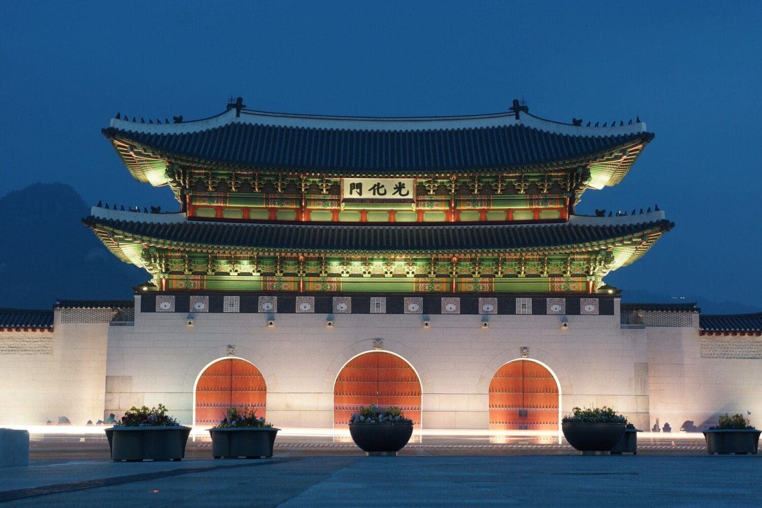 Gwanghwamun Gate illuminated at night, featuring a multi-tiered roof, ornate eaves, and three arched wooden gates on a stone base.