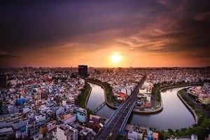 Aerial view of a city at sunset, showing a dense urban area with buildings, winding rivers, and a bridge under an orange and purple sky.