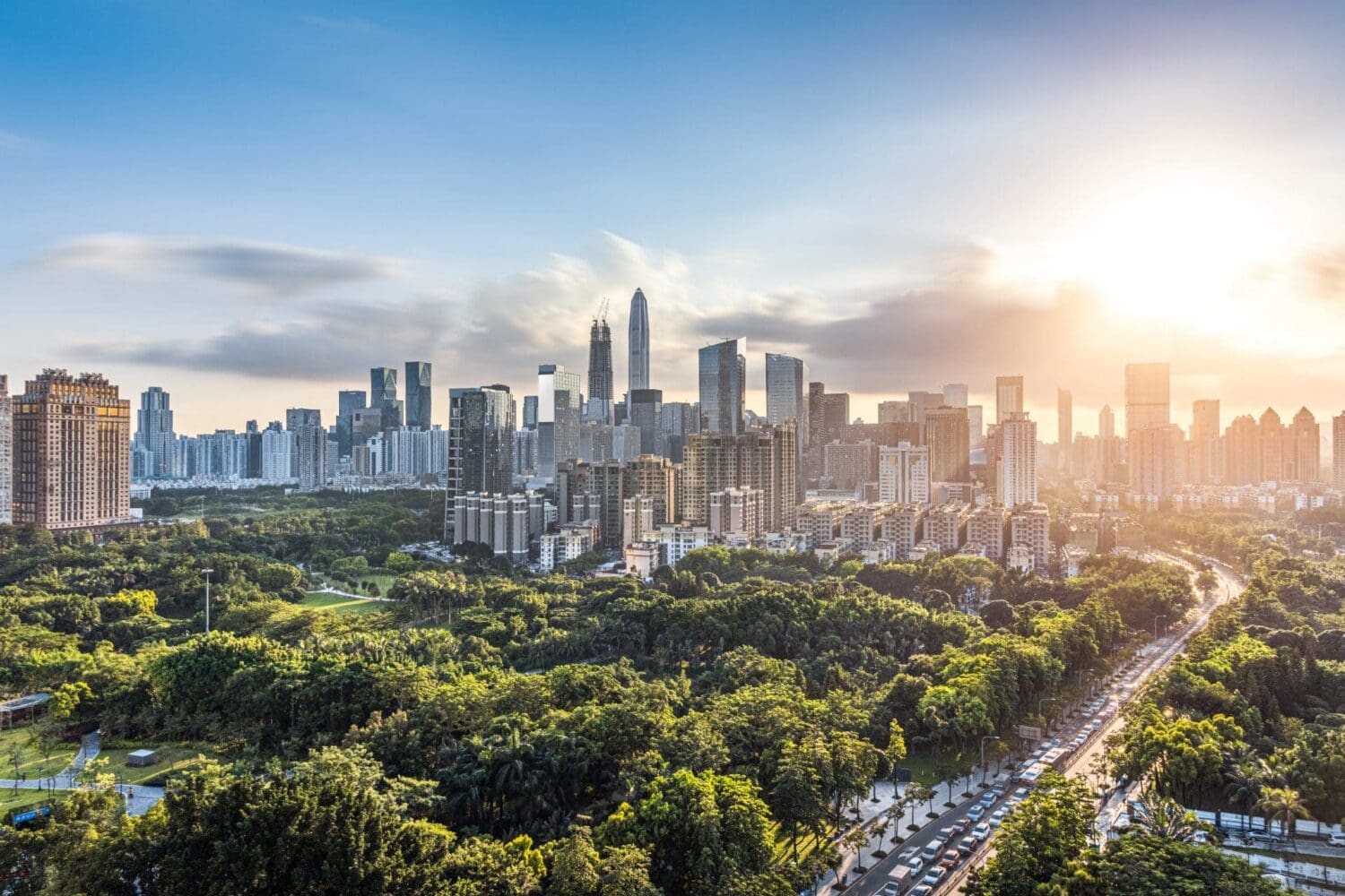 Aerial view of a city at golden hour, showing a skyline of modern skyscrapers, a large green park, and a busy road with cars.