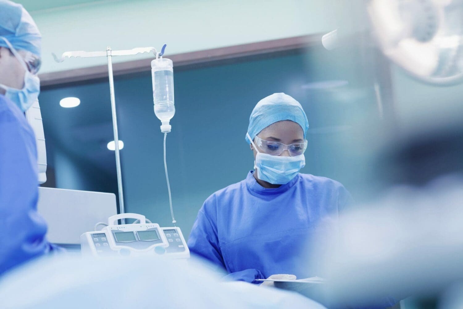 Two medical professionals in blue scrubs, caps, masks, and safety glasses in an operating room. An IV bag and medical equipment are visible.