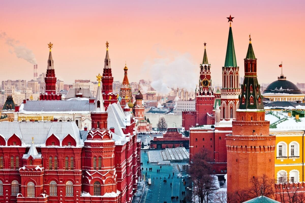 Aerial view of Moscow's Red Square and Kremlin in winter, featuring red brick buildings, snow-covered roofs, and green-spired towers with red stars under a pink and orange sky.