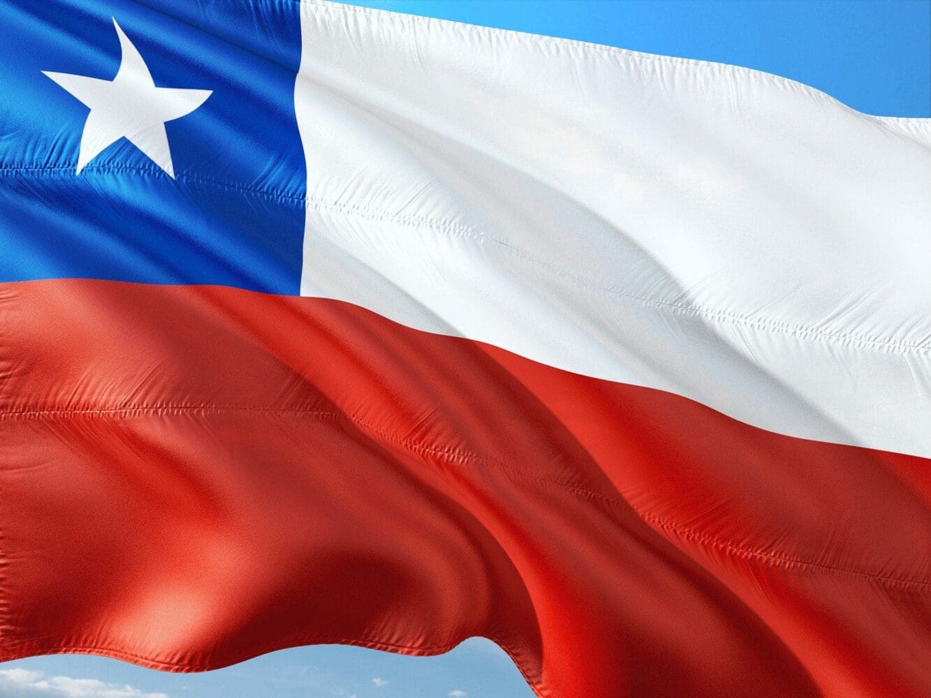 The Chilean flag, featuring a white star on a blue square in the upper left, with a white stripe above a red stripe, waving against a blue sky.