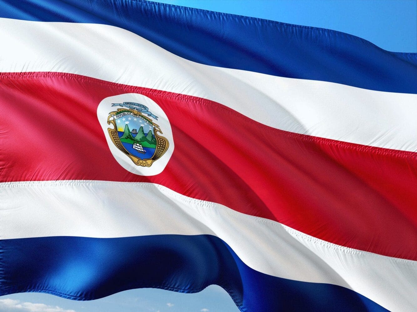 The national flag of Costa Rica waving, showing blue, white, and red horizontal stripes with the national coat of arms on the central red stripe.