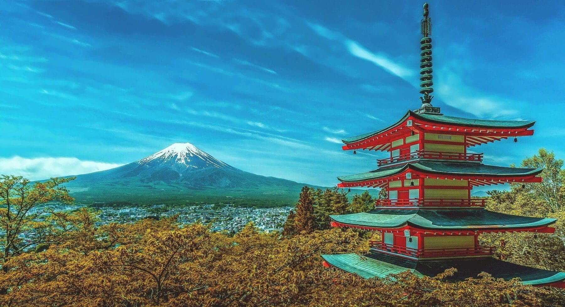 A red and green pagoda with a tall spire stands on the right, surrounded by golden-brown trees. In the background, a snow-capped Mount Fuji rises above a town, all under a bright blue sky with wispy clouds.