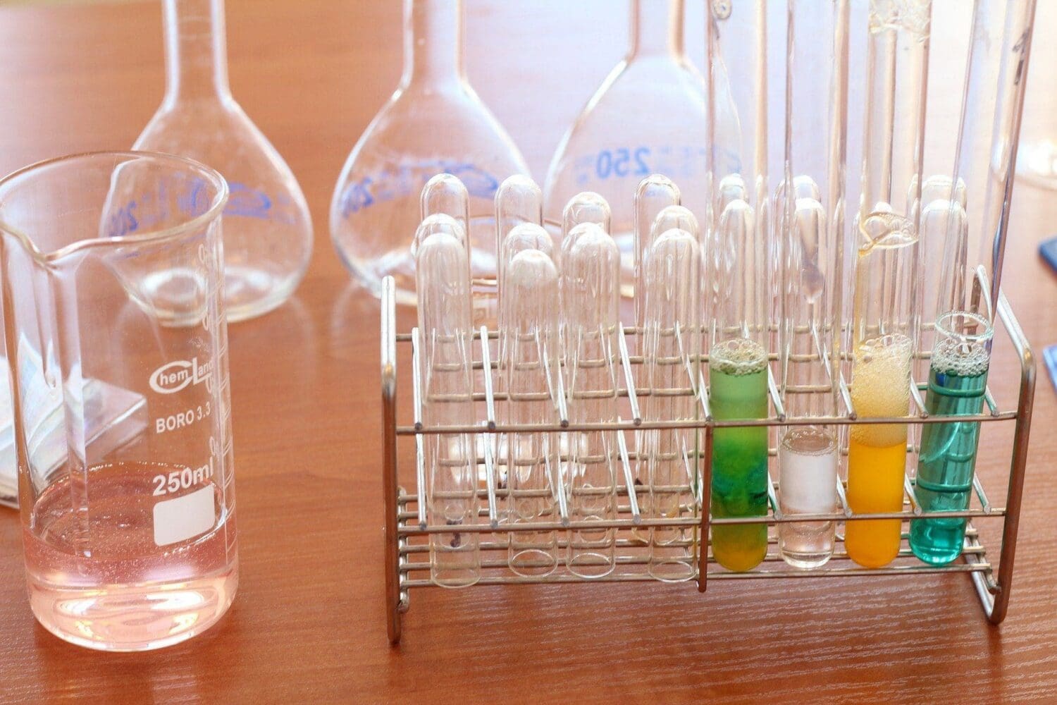 A laboratory setup with a beaker containing pink liquid, several flasks, and a metal rack holding numerous test tubes. Four test tubes on the right contain foamy liquids in green, clear, yellow-orange, and blue-teal colors.