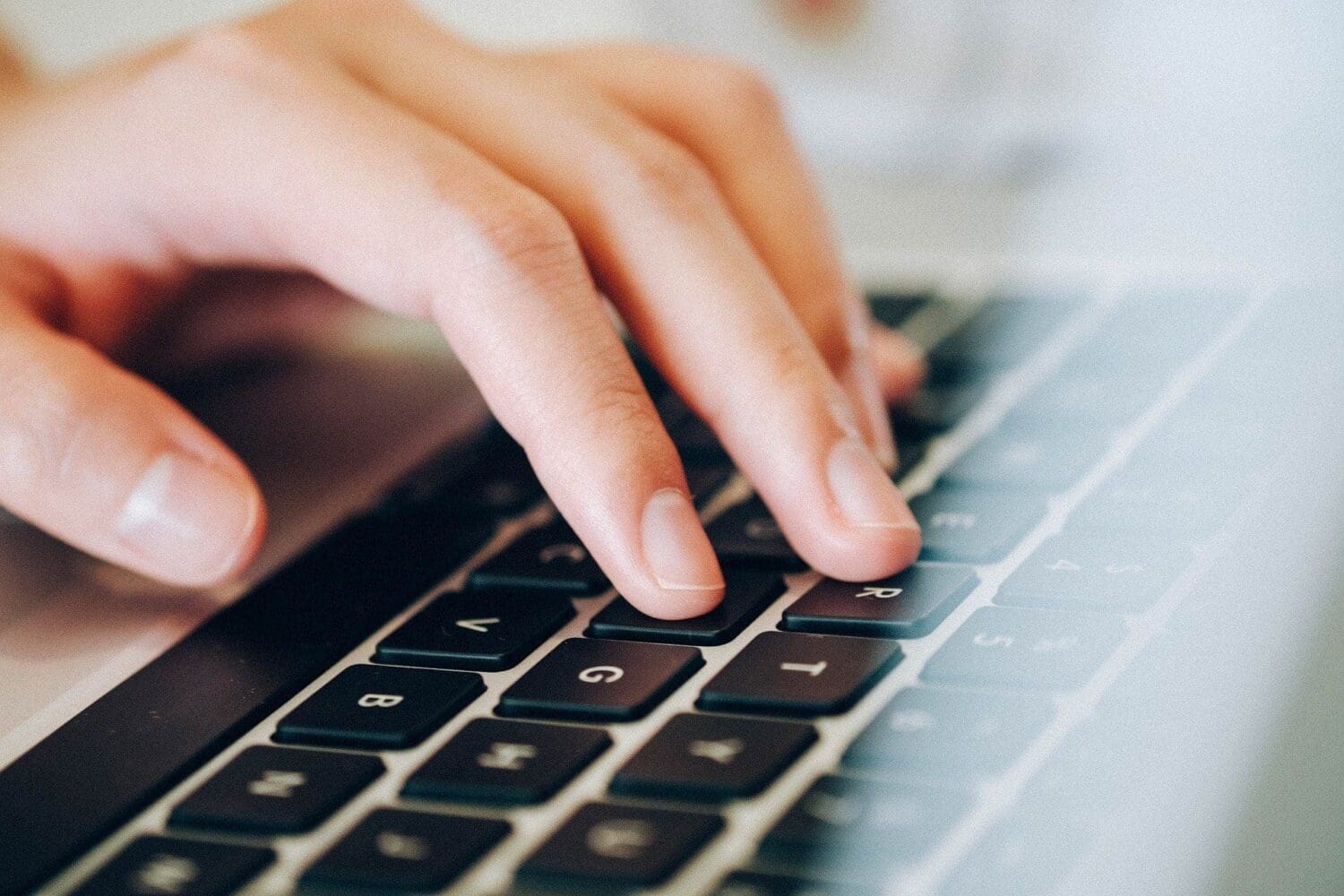Close-up of a hand with light skin tone typing on a black laptop keyboard, with fingers resting on the G, H, and J keys.