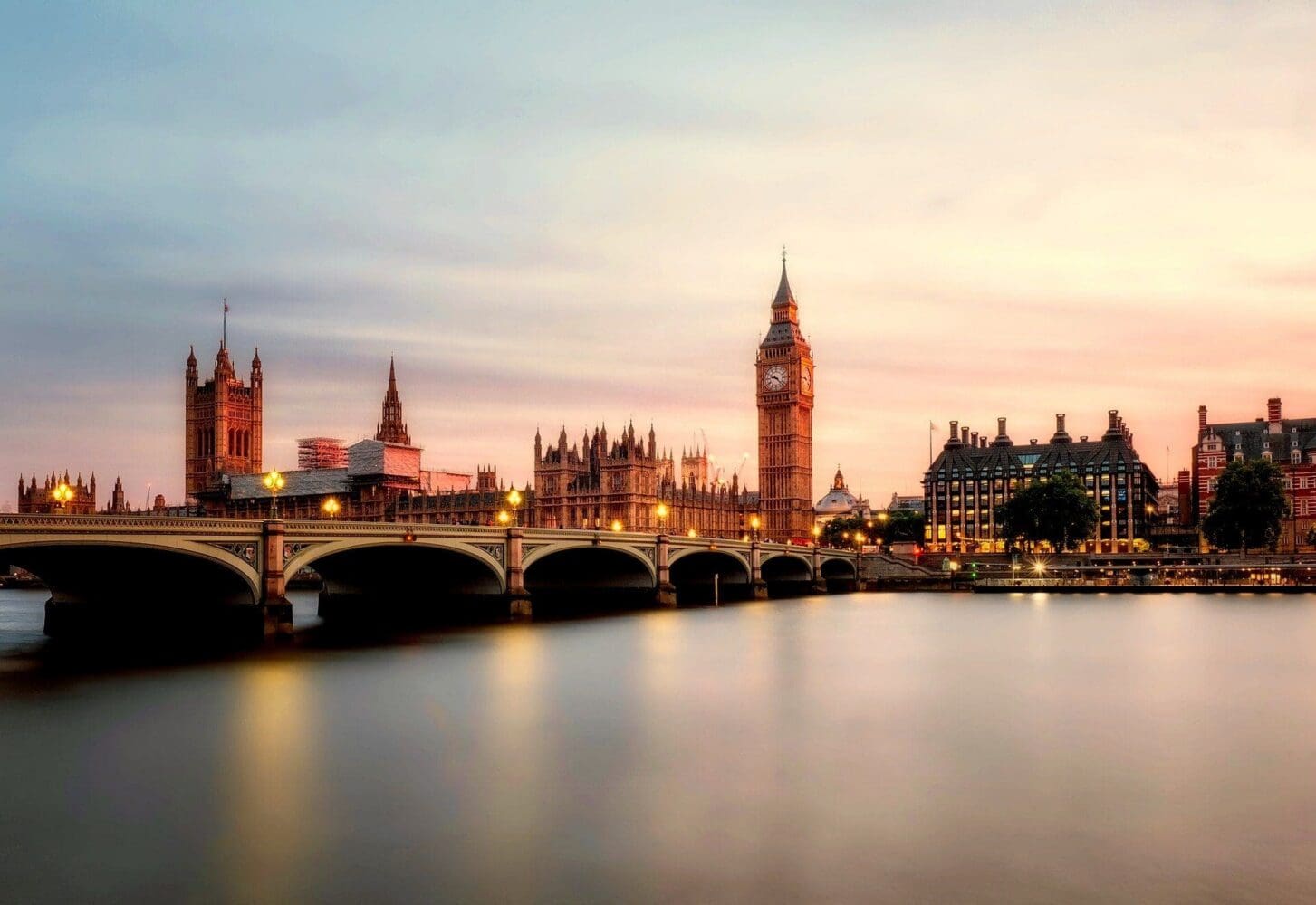 London skyline at dusk with Westminster Bridge, Houses of Parliament, and Big Ben illuminated against a pink and blue sky over the River Thames.