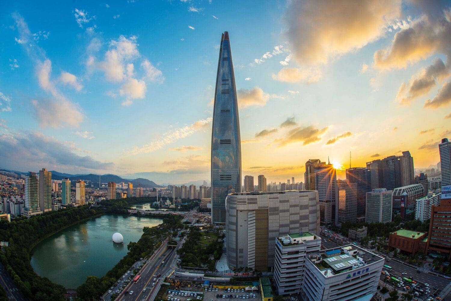 Aerial view of Seoul at sunset, featuring the towering Lotte World Tower, a large lake with a white sphere, and numerous city buildings under a colorful sky.
