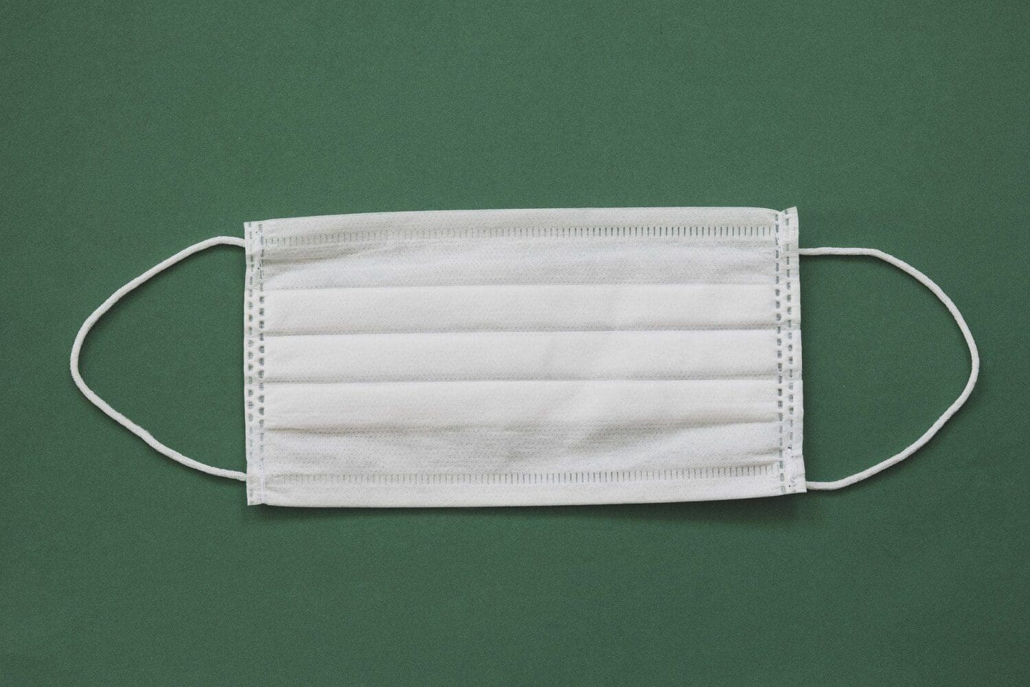 A white disposable face mask with ear loops on a dark green background.