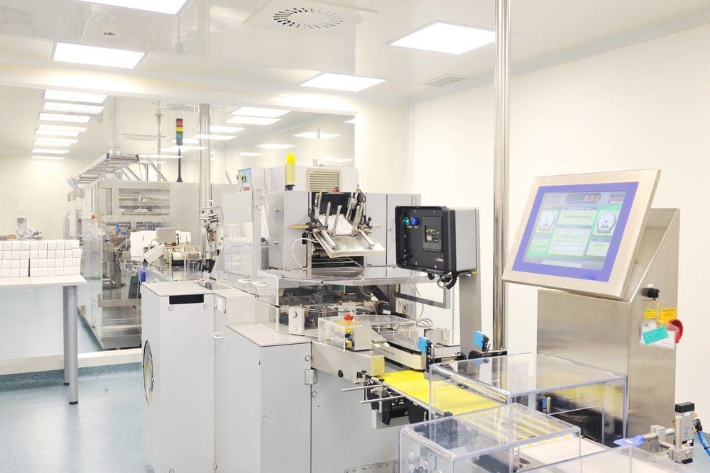 Automated packaging machinery in a cleanroom, featuring conveyor belts, control screens, and stacked white boxes.