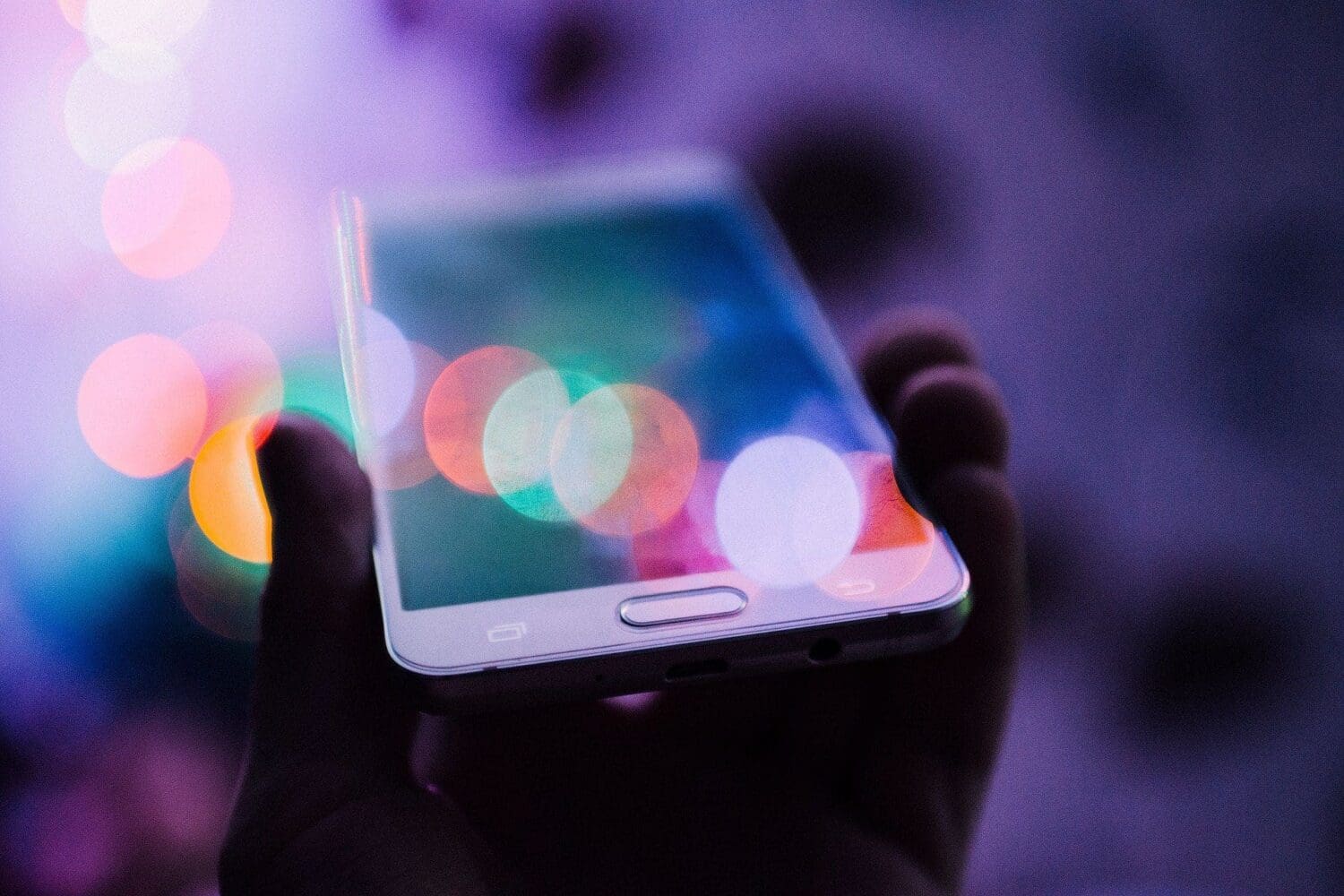 A hand holds a smartphone, its screen reflecting colorful, out-of-focus bokeh lights in purple, pink, orange, green, and blue.