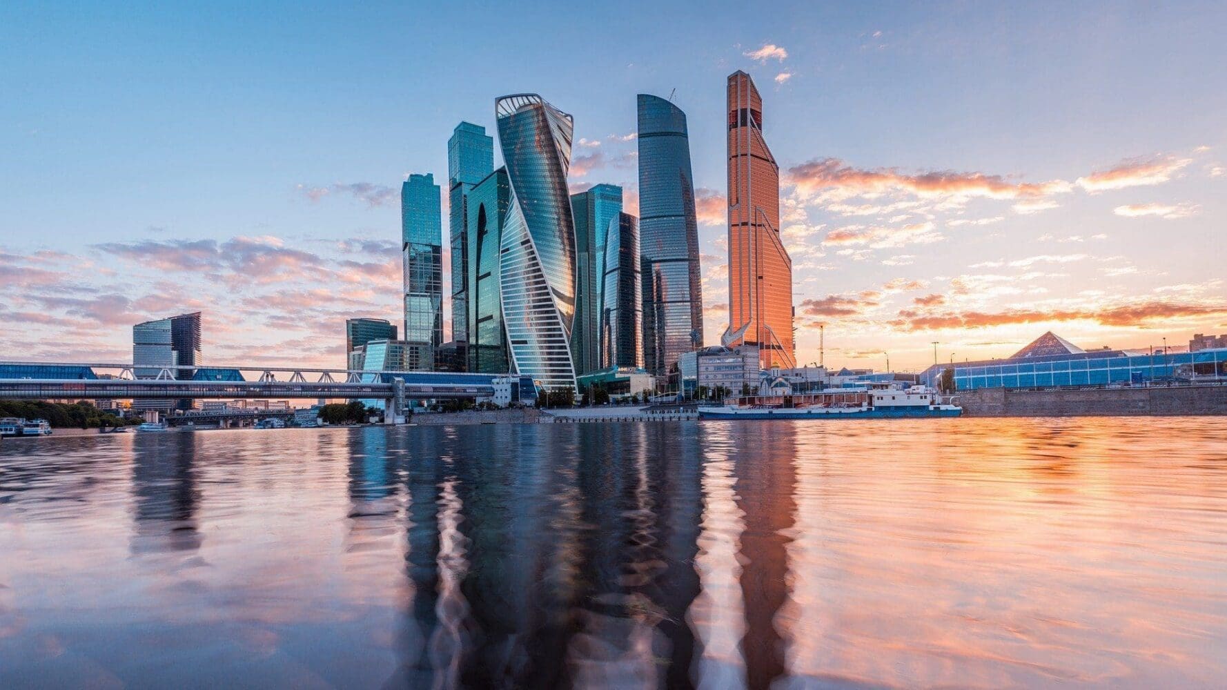 A modern city skyline with multiple skyscrapers reflected in a river under a colorful sunset sky.
