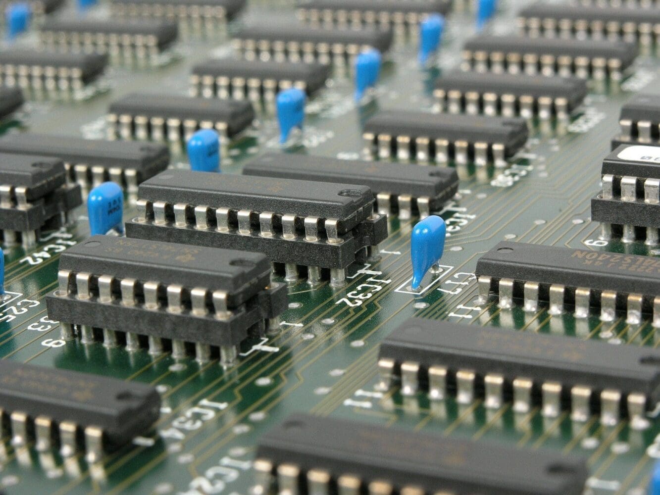 Close-up of a green circuit board with many black integrated circuits and blue capacitors.