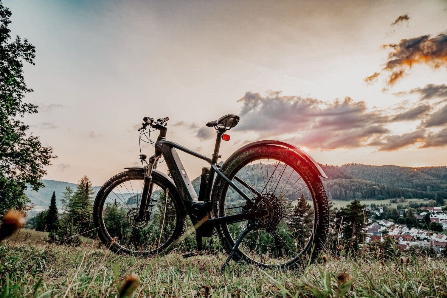 An electric mountain bike stands on a grassy hill, overlooking a village and forested hills under a sunset sky.