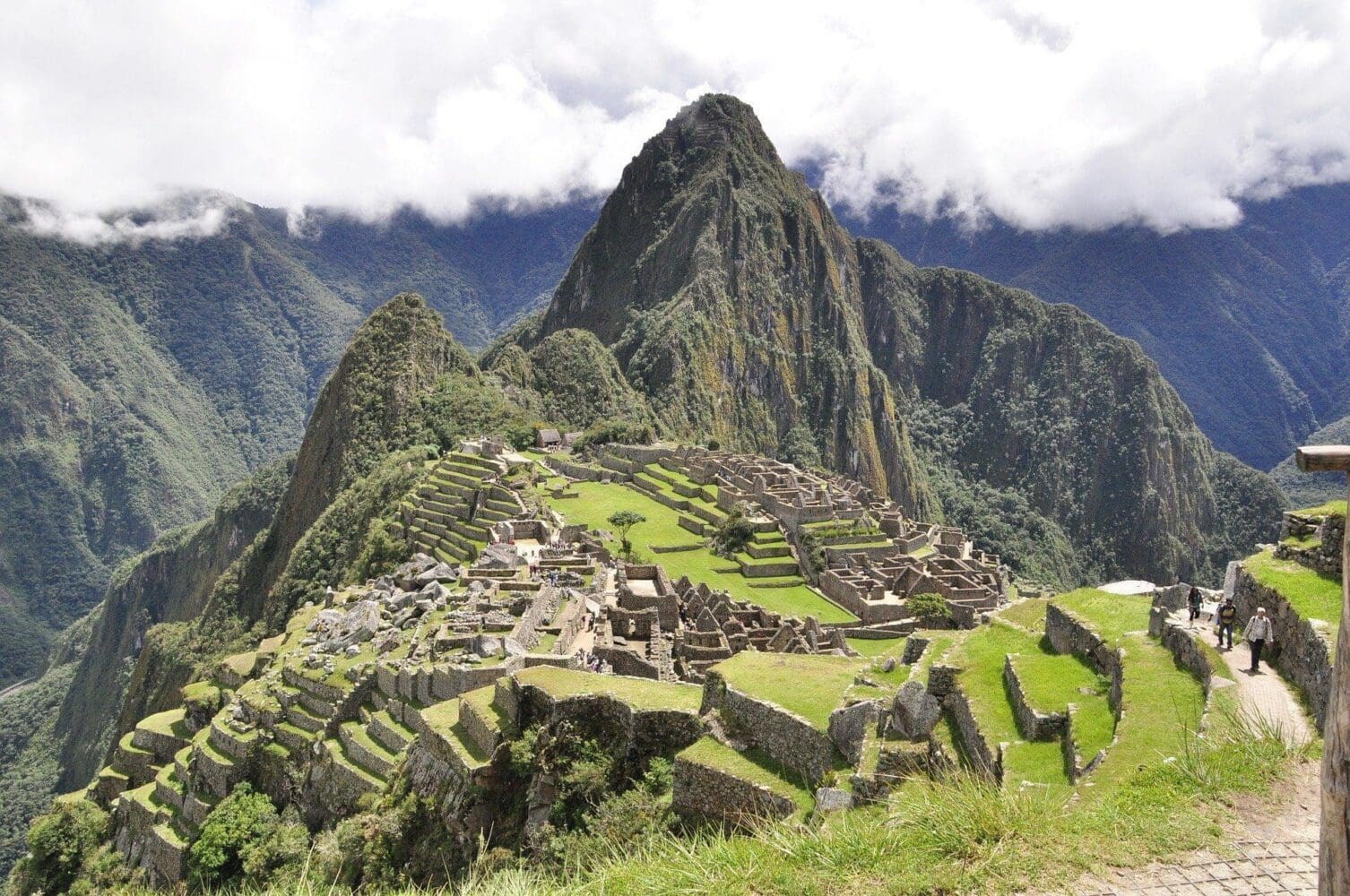 Panoramic view of Machu Picchu, showing ancient stone ruins, agricultural terraces, and the towering Huayna Picchu mountain under a cloudy sky.