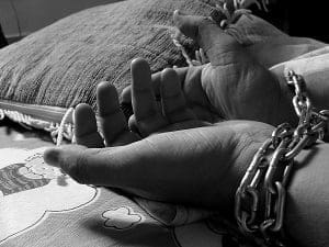 Black and white image of a person's hands, with one wrist bound by a metal chain, resting on a patterned pillow.
