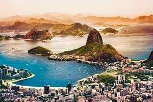 Aerial view of a coastal city with a large, green, sugarloaf-shaped mountain in a bay, surrounded by urban development and distant mountains.