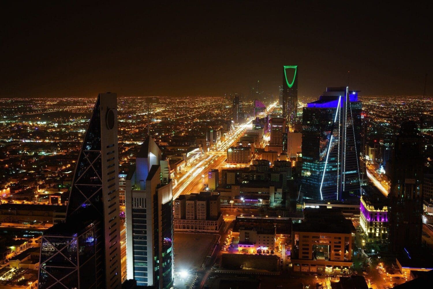 Night aerial view of a brightly lit city skyline with skyscrapers, including the green-lit Kingdom Centre, and light trails from traffic on a highway.