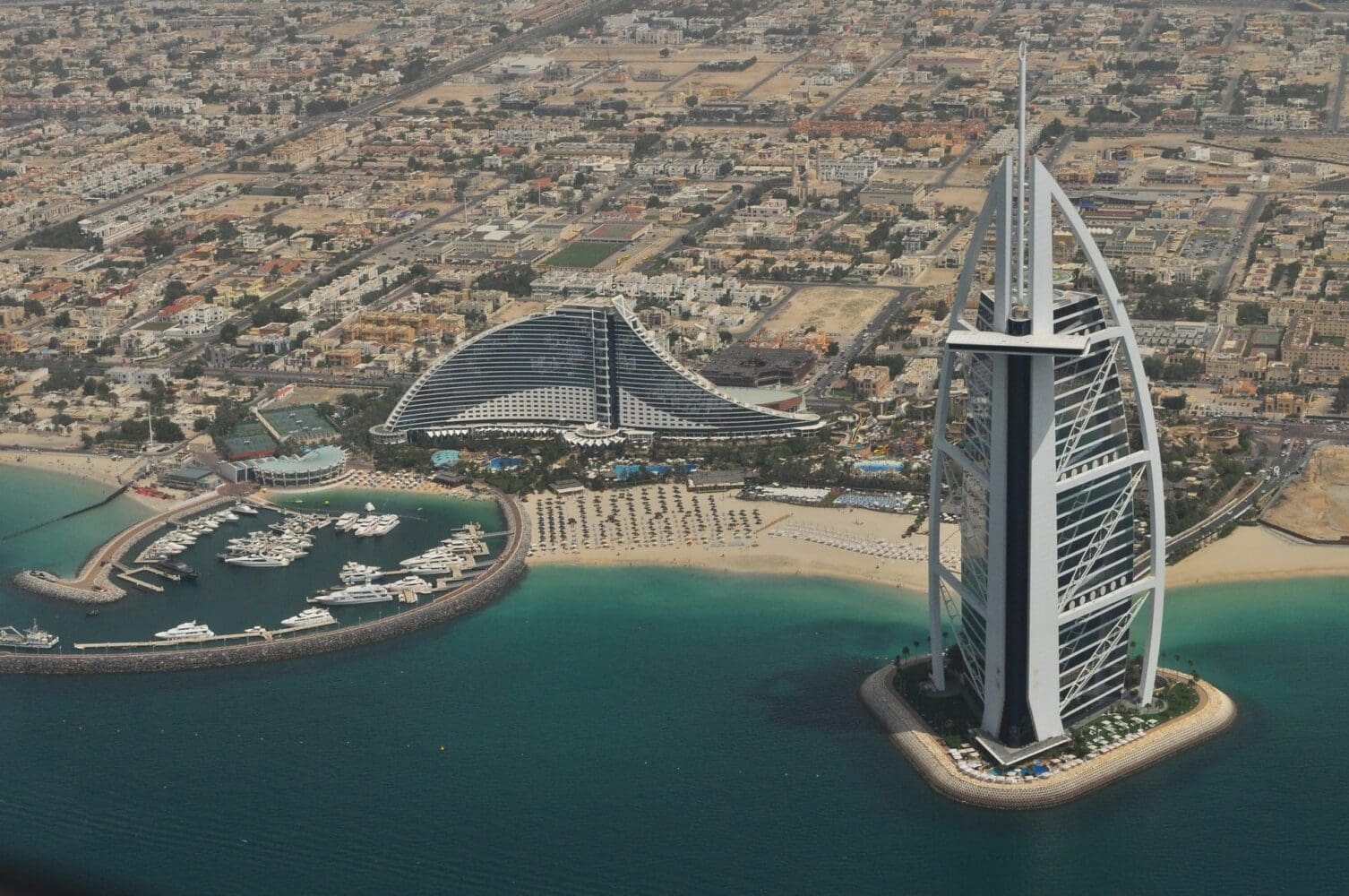 Aerial view of Dubai showing the sail-shaped Burj Al Arab and wave-shaped Jumeirah Beach Hotel, a marina with yachts, sandy beaches, and the city skyline.