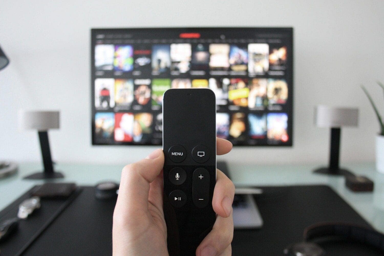 A hand holds a black remote control in front of a TV screen displaying a streaming service interface with movie thumbnails.