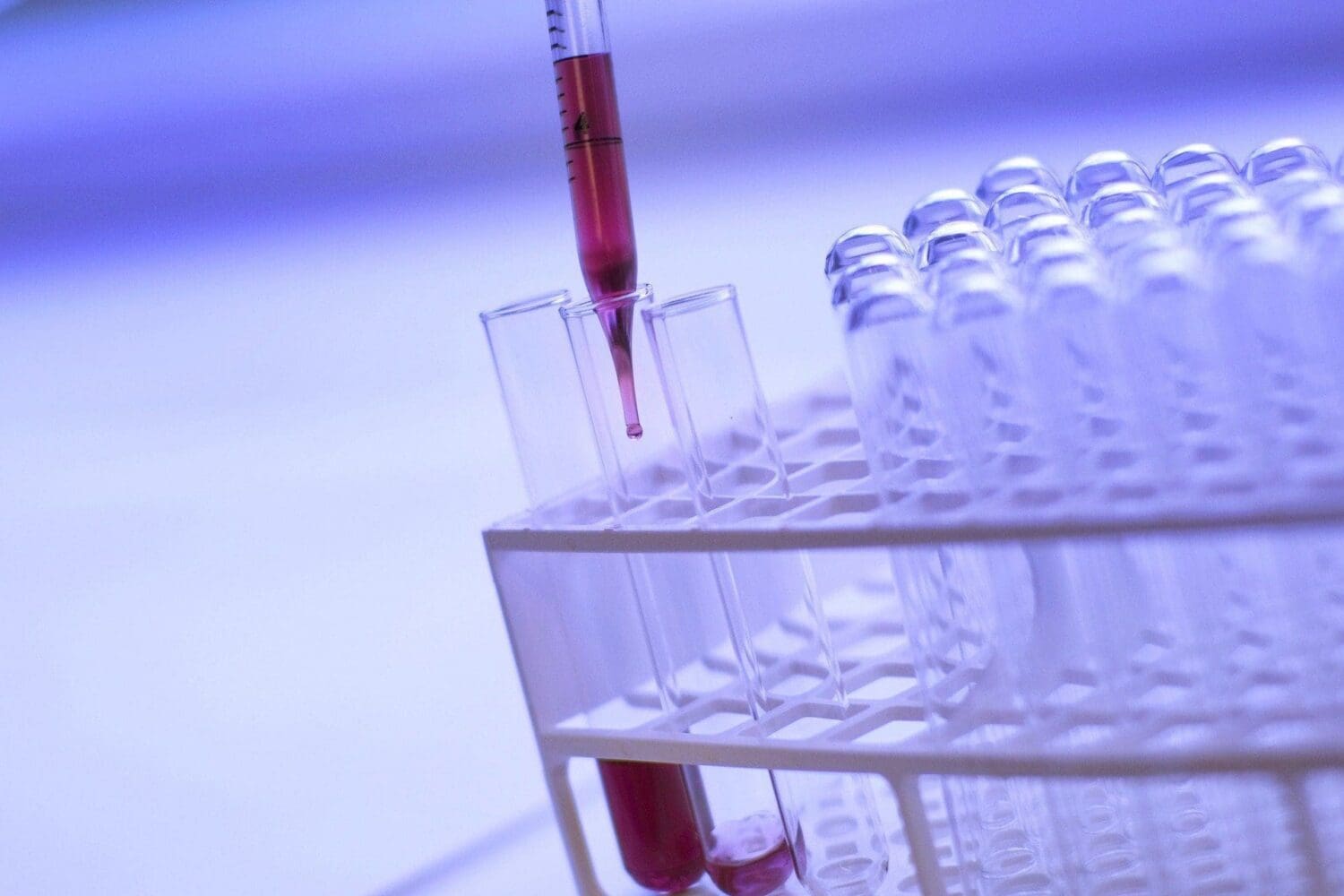 A close-up of a pipette dispensing red liquid into a clear test tube in a white rack, with other test tubes visible.