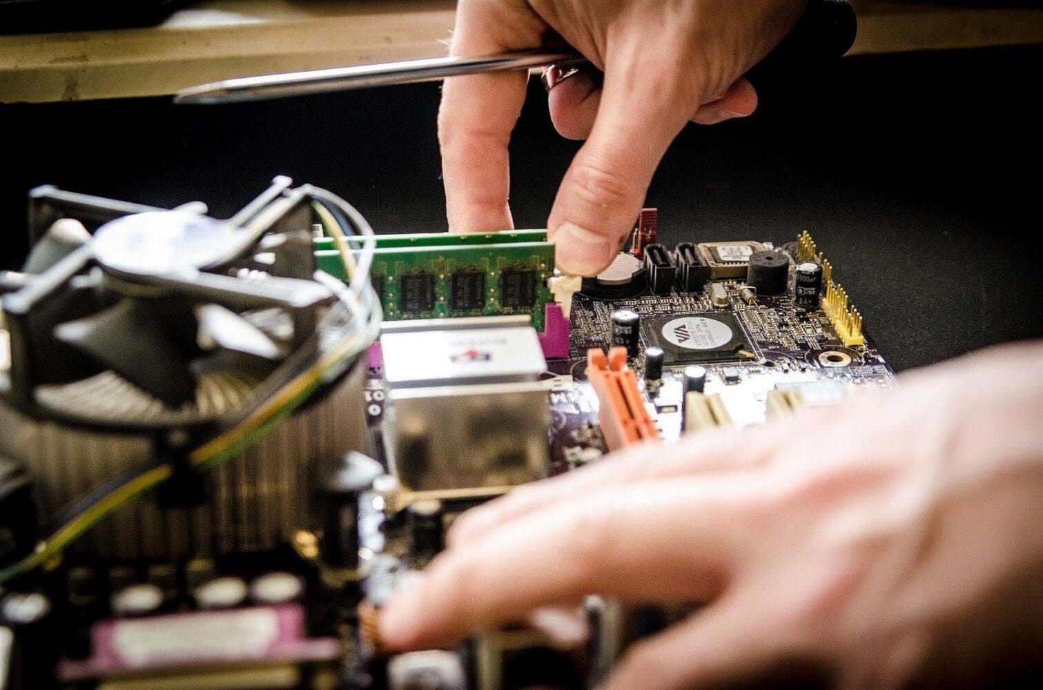 Close-up of hands installing a green RAM module into a computer motherboard with a screwdriver.