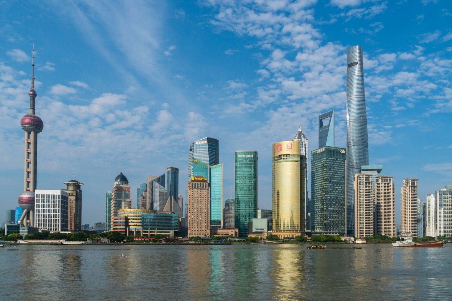 A panoramic view of the Shanghai skyline across a river, showing the Oriental Pearl TV Tower, Shanghai Tower, and other skyscrapers under a blue sky with clouds.