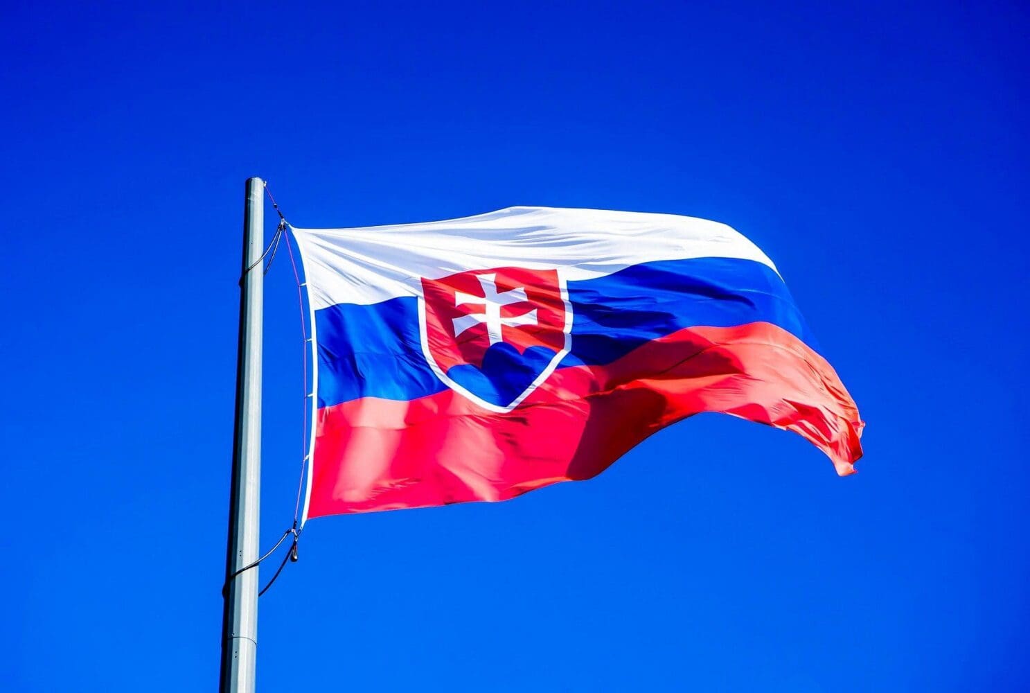 The flag of Slovakia, with white, blue, and red horizontal stripes and the national emblem, waving against a clear blue sky.