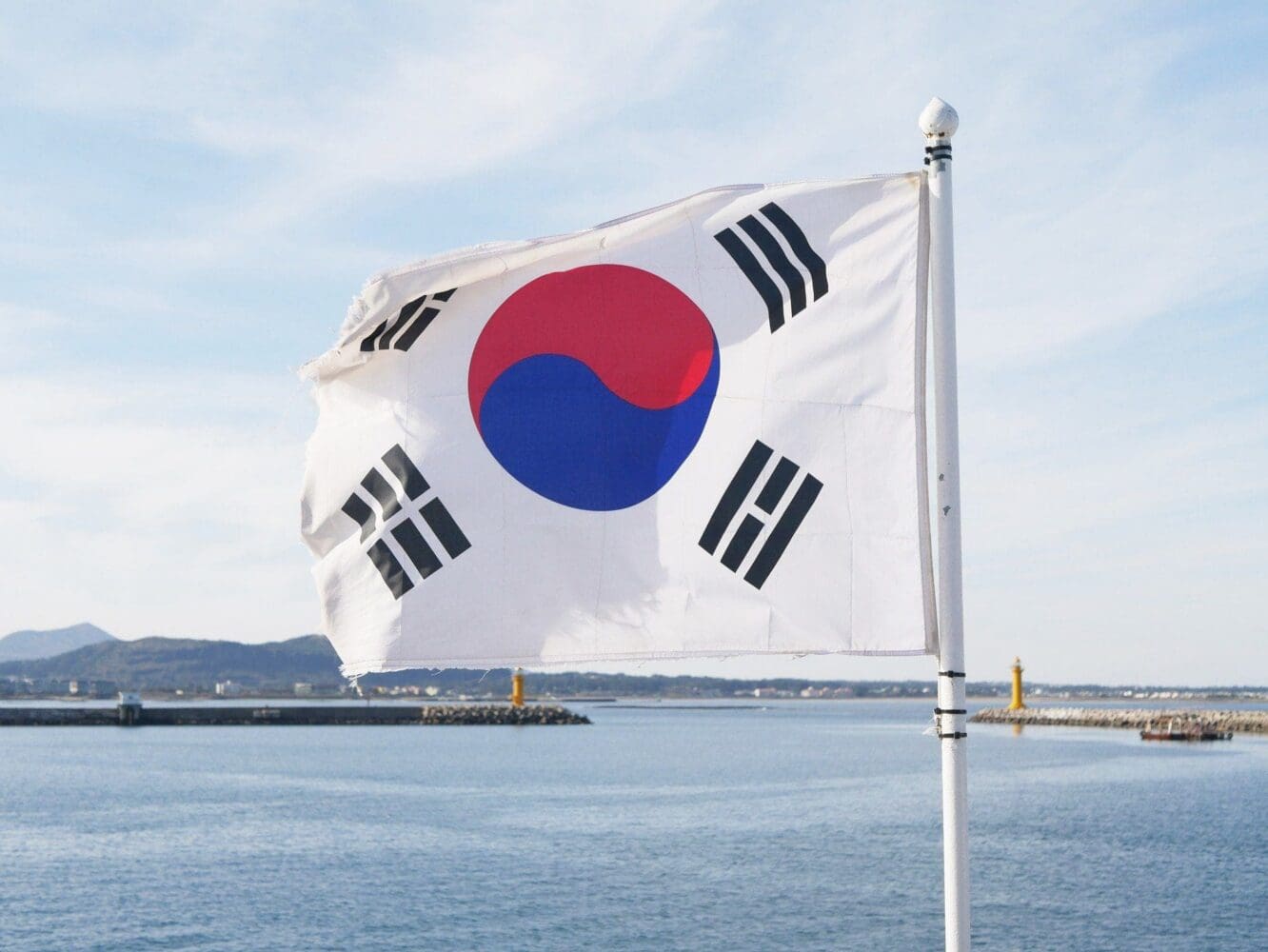 The South Korean flag, white with a red and blue circle and black trigrams, waves above a blue sea with distant lighthouses and land.