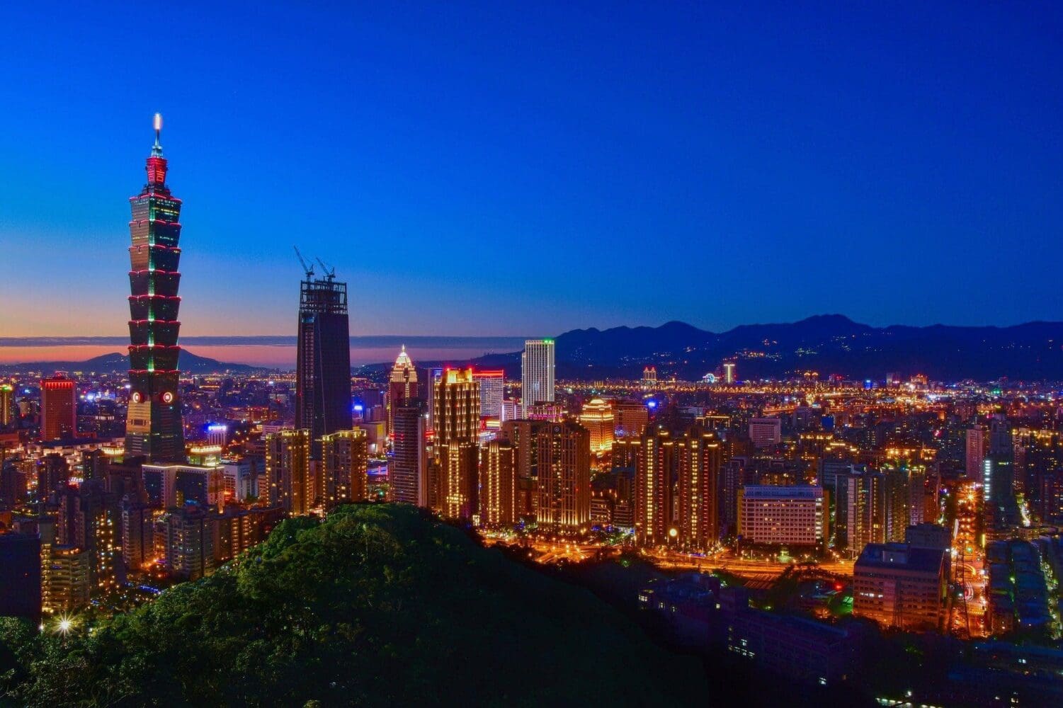 Panoramic view of Taipei city skyline at twilight, featuring the illuminated Taipei 101 skyscraper, other glowing buildings, and mountains in the background.