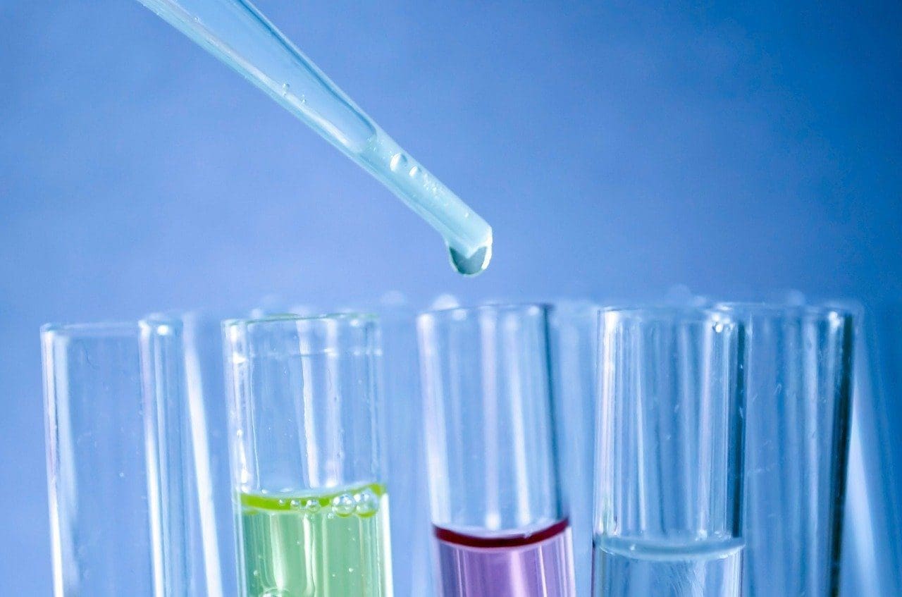 A dropper with a suspended drop above test tubes containing green, purple, and clear liquids against a blue background.