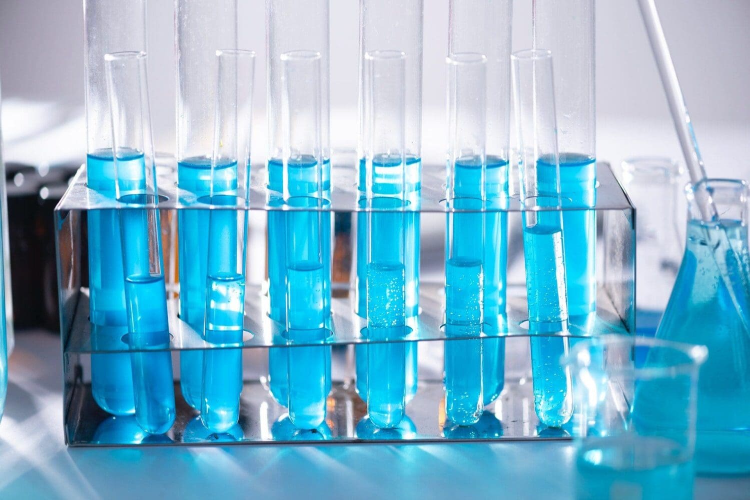 Close-up of glass test tubes in a metal rack, filled with bright blue liquid. A conical flask and beaker with blue liquid are in the background.