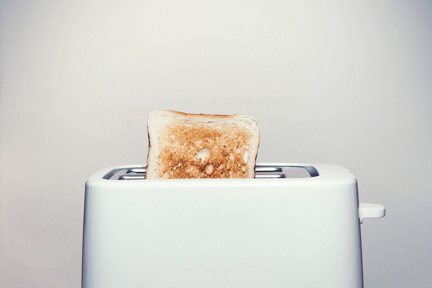 A slice of golden brown toast popping out of a white toaster against a light background.