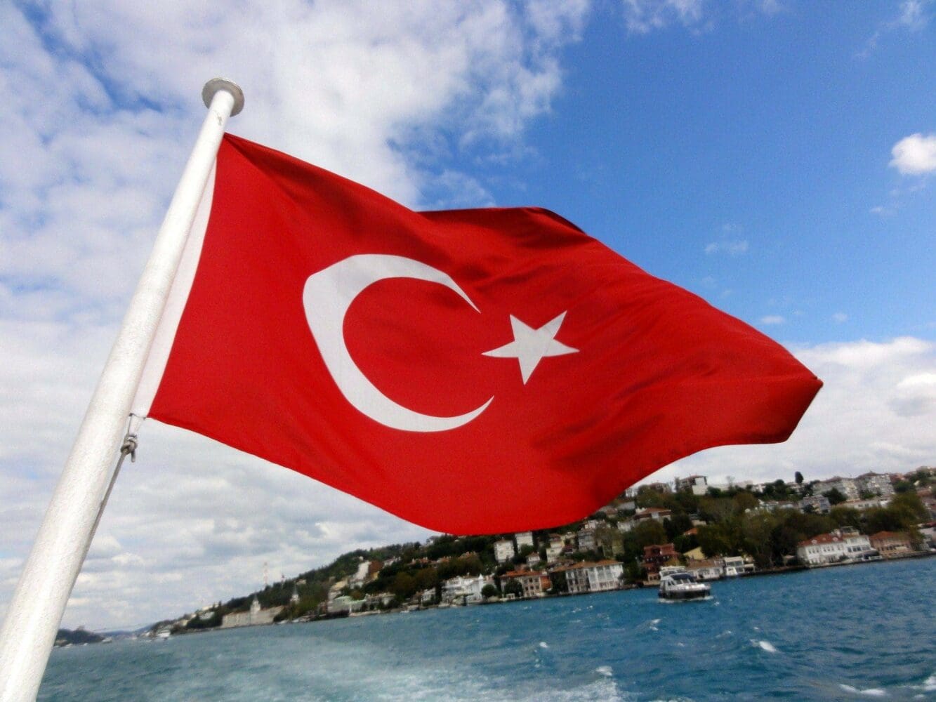 The red Turkish flag with a white crescent and star waves from a flagpole, with a blue waterway and a city-lined coast in the background.
