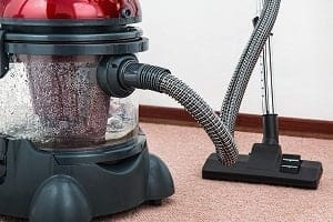 A red and black vacuum cleaner with a transparent water tank containing dirty liquid, a hose, and a floor nozzle on a light brown carpet.