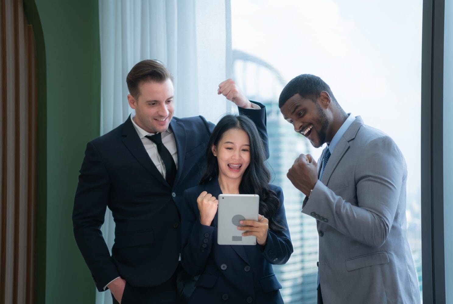 Three diverse business professionals, two men and one woman, celebrating excitedly while looking at a tablet.