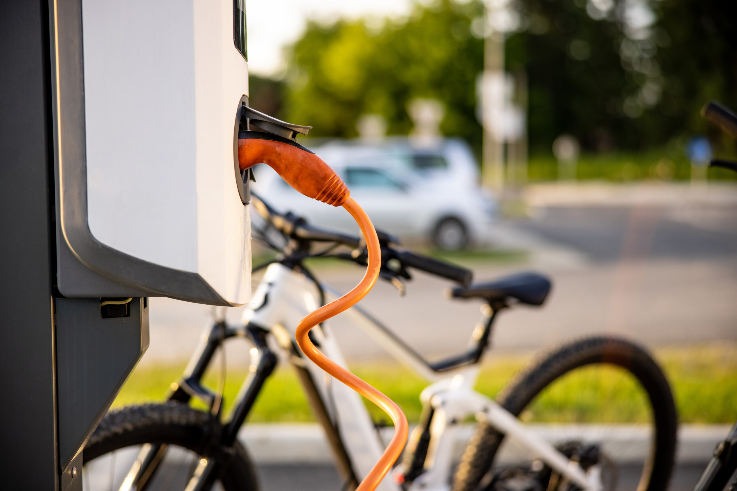 Close-up of an orange charging cable plugged into a white charging station, with a blurred e-bike and car in the background.