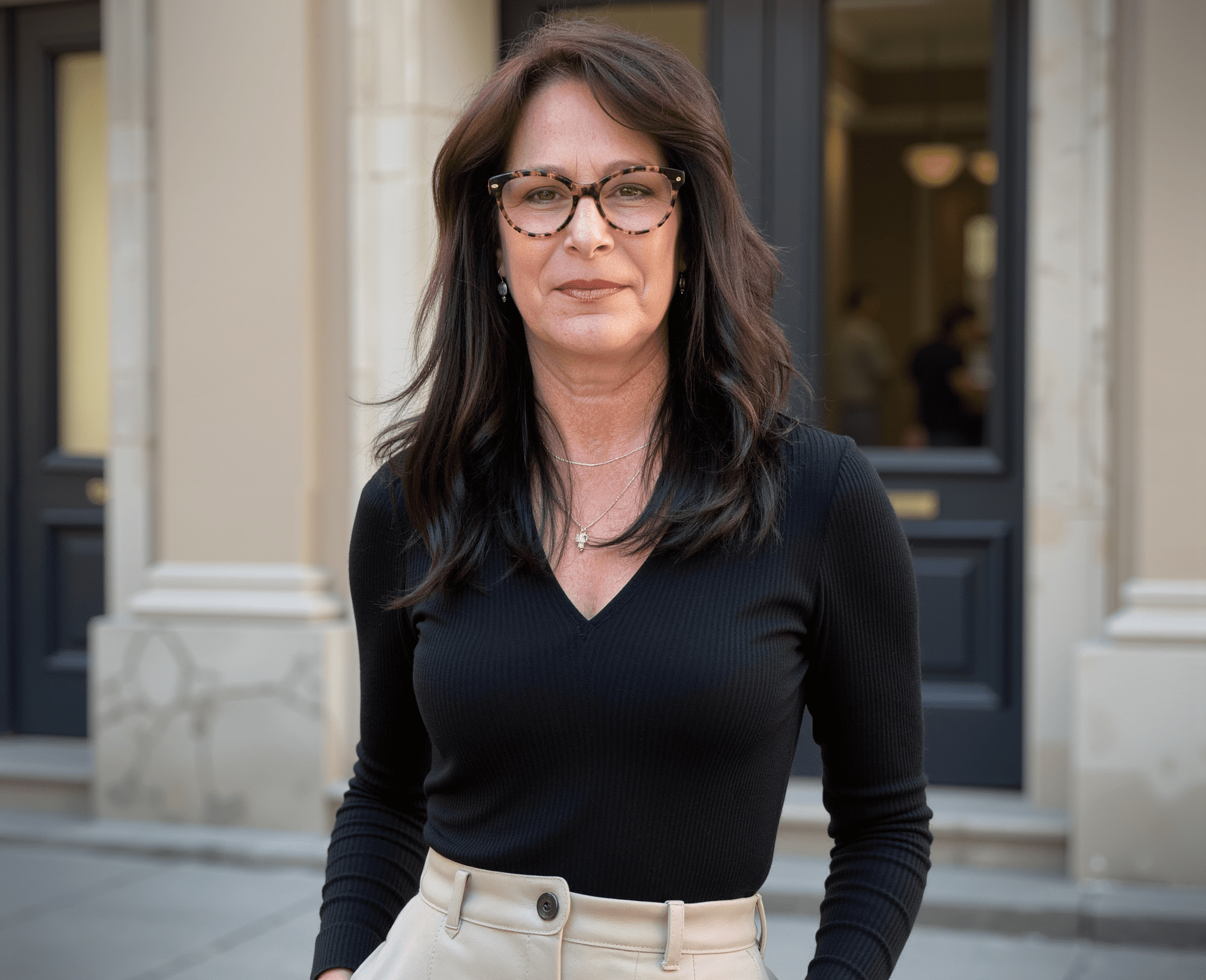 A woman with long brown hair and tortoise-shell glasses smiles faintly at the camera. She wears a black V-neck top and beige pants, with a building in the blurred background.