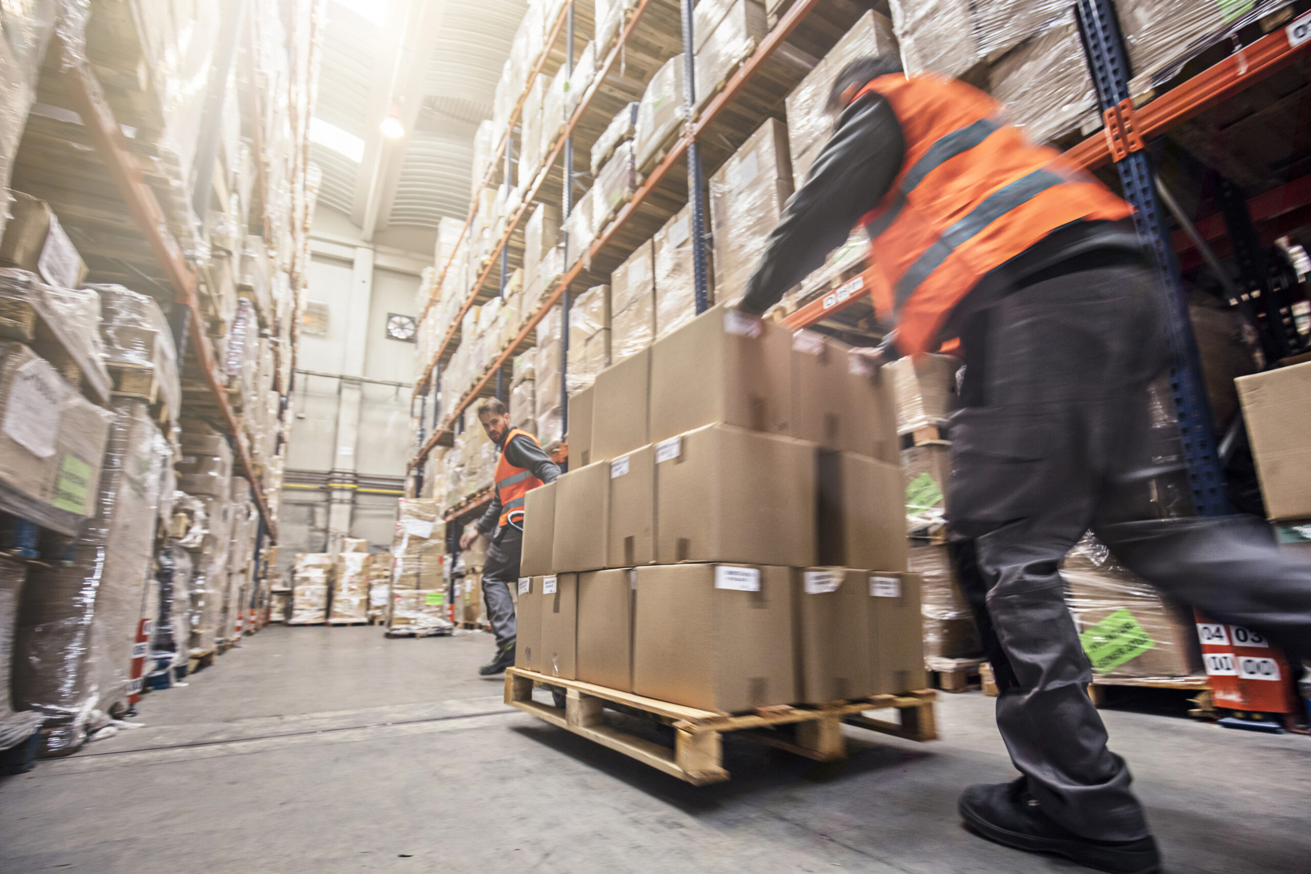 Two warehouse workers in orange vests moving stacks of cardboard boxes on a pallet jack amidst tall shelving units filled with inventory, conveying a sense of busy operations.
