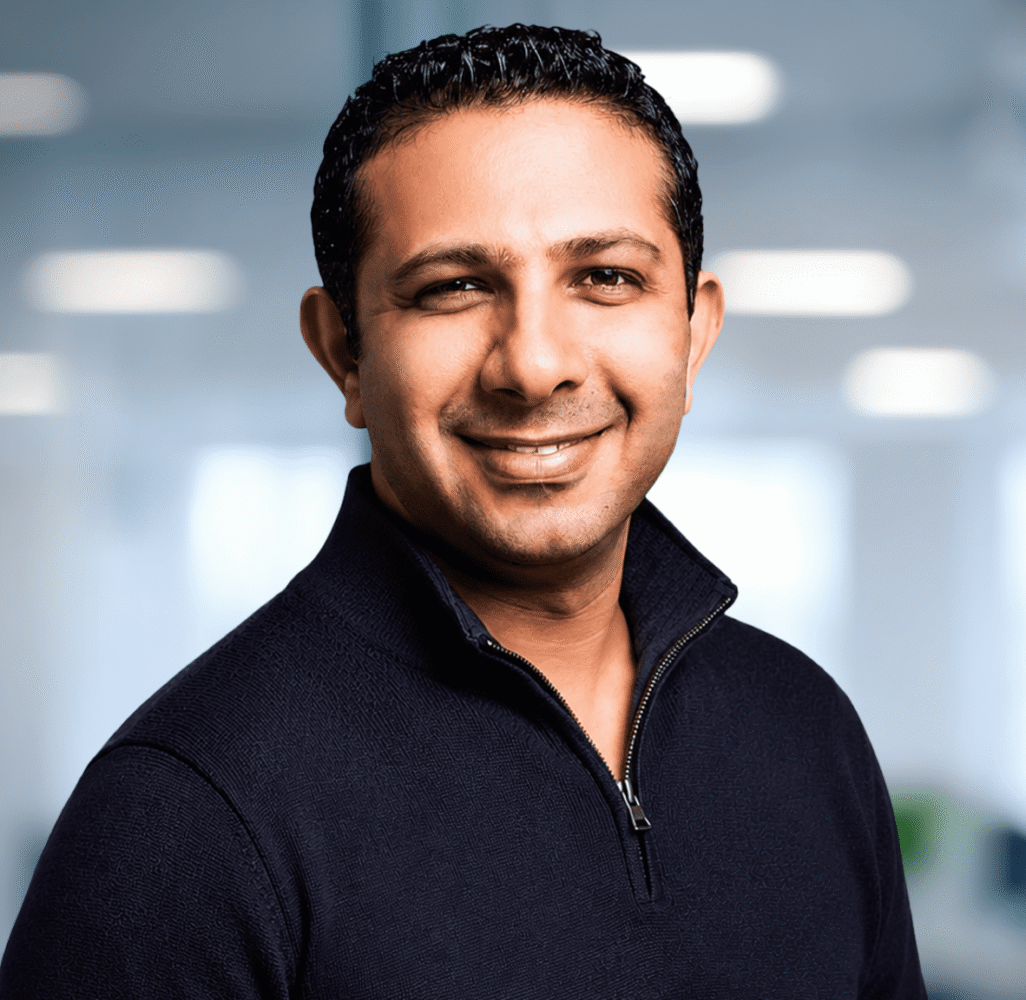 Headshot of a smiling man in a dark quarter-zip sweater against a blurred office background with a plant.