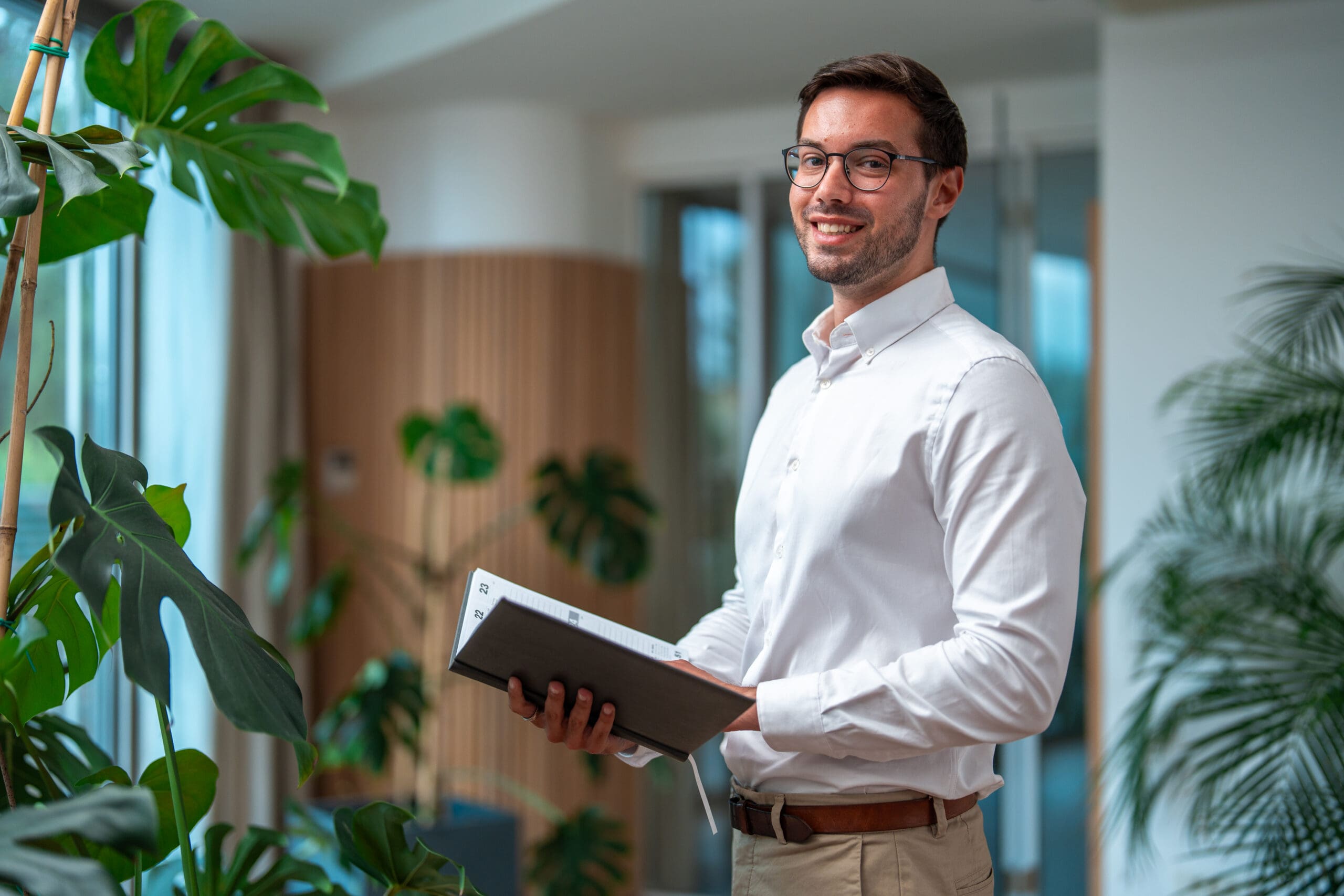 A smiling man in a white shirt and glasses holds an open planner in a modern office with plants.