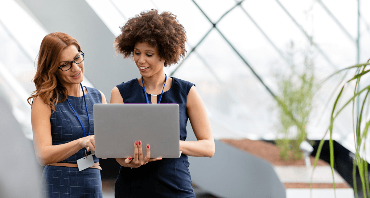 Two women, one with glasses and one with curly hair, smiling and looking at a silver laptop. They are wearing lanyards and standing in a bright, modern building with large windows and plants in the background.