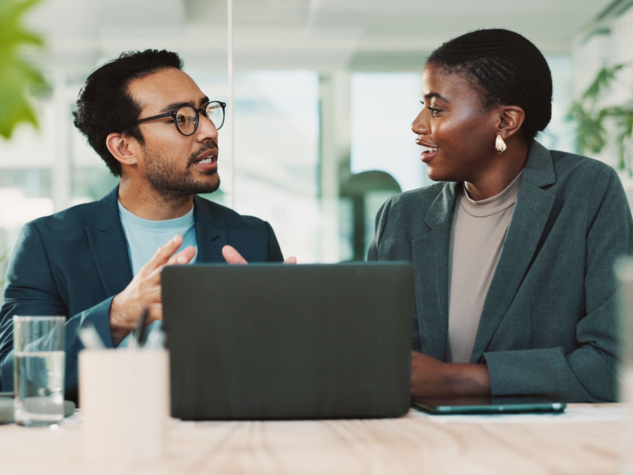 A man and a woman in business attire are seated at a table, facing each other and conversing. A laptop is open between them.