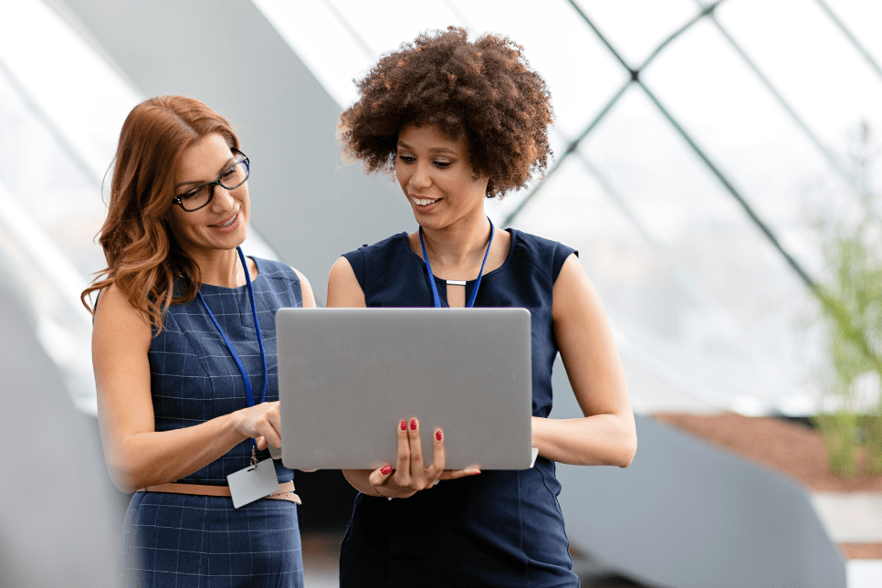 Two women, one with glasses and one with curly hair, smiling and looking at a silver laptop. They are wearing lanyards and standing in a bright, modern building with large windows and plants in the background.